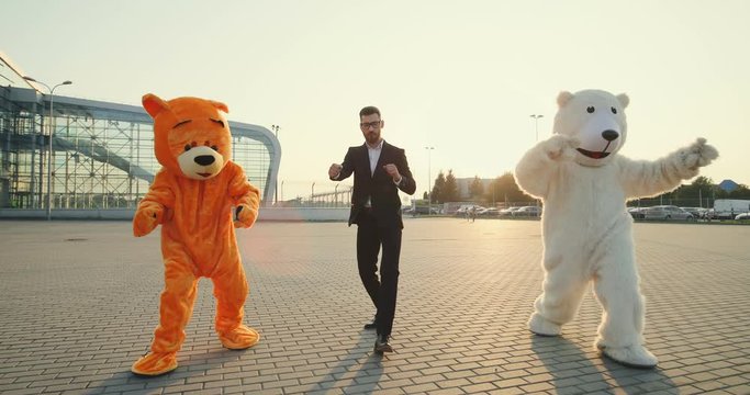 Cheerful Caucasian young handsome businessman having fun, dancing and jumping outdoors the big urban building with two funny growth dolls of bears.