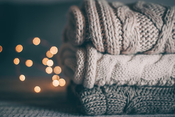 Stack of cozy knitted sweaters and garland lights on wooden background. Autumn-winter concept. Magic, cozy and mood time