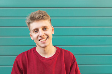 Handsome young man in a red sweatshirt against a turquoise wall looks at the camera and smiles. Emotional attractive man smiles.