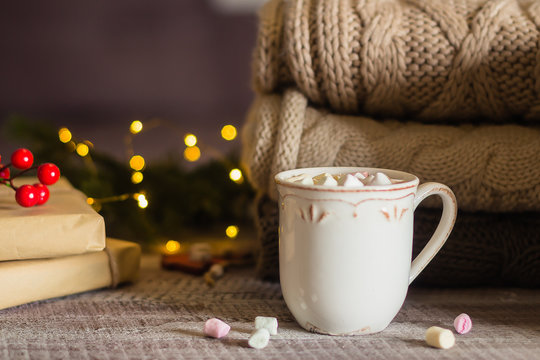 Stack of cozy knitted sweaters and cup of coffee or hot chocolate with marshmallow on wooden background. Autumn-winter concept. Magic, cozy and mood time