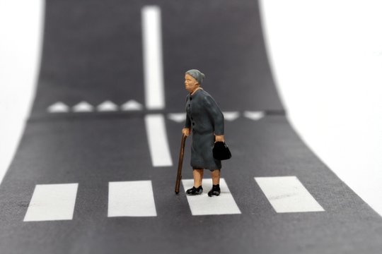 persona anziana in miniatura che attraversa la strada sulle strisce pedonali aiutandosi col bastone