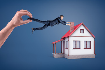 A businessman tries to hold onto a chimney of a small house while he is dragged away by a giant hand.