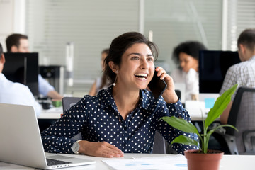 Excited hindu business woman happy to hear good news talking on cell phone in office, smiling...