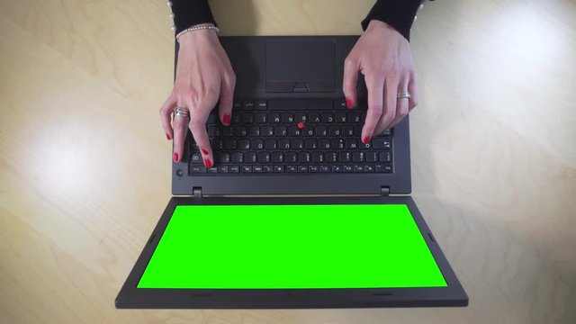 female hands working on a laptop with green screen over a wooden table. Top view