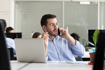 Male entrepreneur making call consulting client speaking by cell near laptop in office, broker...