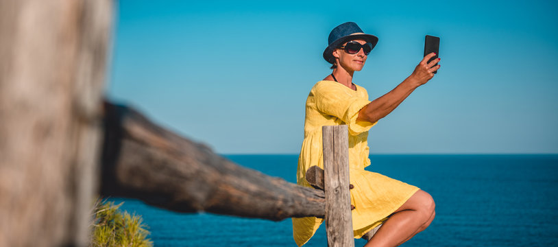 Woman sitting on a wooden fence by the sea and taking selfie