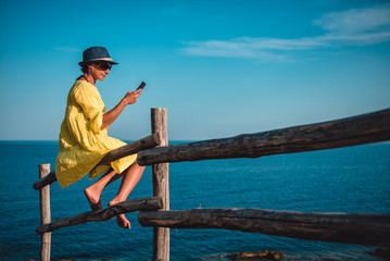 Naklejka premium Woman sitting on a wooden fence by the sea and using smart phone