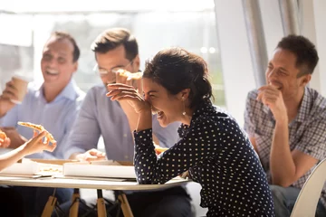 Fotobehang Indian woman laughing at funny joke eating pizza with diverse coworkers in office, friendly work team enjoying positive emotions and lunch together, happy colleagues staff group having fun at break © fizkes