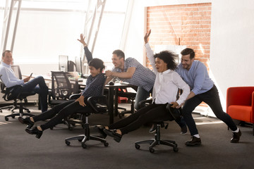 Fototapeta na wymiar Carefree excited diverse workers having fun riding oh chairs celebrating friday together, happy employees enjoy funny competition laughing together feel great at work break, friendly office team game