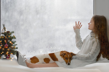 Cute little girl and her puppy jack russell sitting by the window