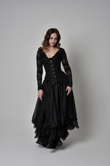 full length portrait of brunette girl wearing long black gown with corset. standing pose on grey studio background.