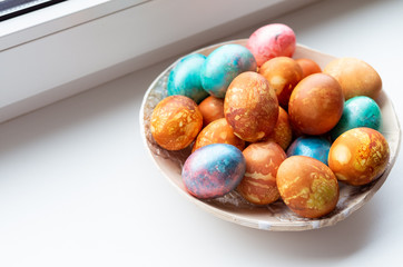 Closeup of colourful dyed Easter eggs in a ceramic bowl on a white window sill.