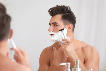 Young man shaving near mirror in bathroom