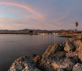 Barnacle covered rocks line the entrance to the harbor with shining lamps and house lights illuminate the city hillside at dusk.