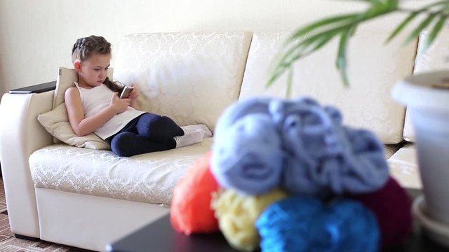A girl with a mobile phone on the couch. On the table there are threads for knitting in different colors.