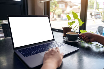 Fototapeta premium Mockup image of a woman's hands using laptop with blank white desktop screen while drinking hot coffee on wooden table in cafe