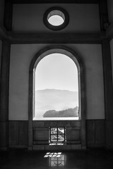 Black and white picture of a landscape with mountains, mist and a lake. The view is through an old open window with an arch and a little round opening above the window 