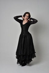 full length portrait of brunette girl wearing long black gown with corset. standing pose on grey studio background.