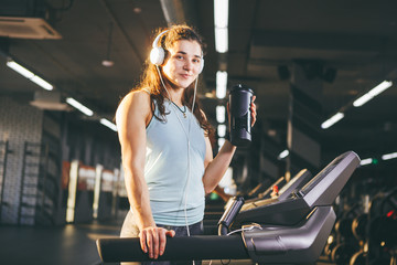 Sports nutrition and exercise. beautiful woman holds protein in her hand in bottles, shaker, listens music in headphones on head in gym running on treadmill, at hands of sports watch,in sunny weather