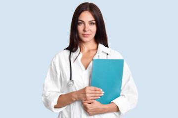 Waist up shot of female doctor holds blue folder, ready to examine patients, wears medical uniform, looks seriously at camera, isolated over light blue studio wall. Medicine and heath care concept