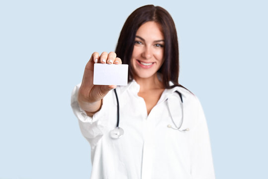 Positive doctor or nurse with pleasant appearance, dressed in medical robe, holds blank card, focus on empty space for your advertising content, isolated over light blue background, works in hospital