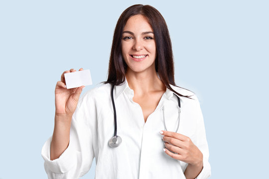 Cheerful young female doctor dressed in uniform, has stethoscope on neck, holds blank empty card for your advertisement or text, poses against blue background. Health care and medicine concept