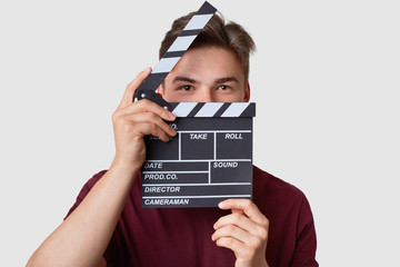 Headshot of handsome man hides face with clapper board, looks seriously at camera, dressed in...