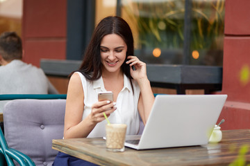 Pleased dark haired woman holds modern cell phone, reads positive news on internet website, drinks cocktail in outdoor cafeteria, sits in front of opened laptop, connected to wireless internet