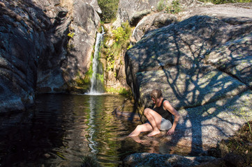 Caucasian woman sitting on a big stone, in the summer, near a little lake with a waterfall with her feet in the water