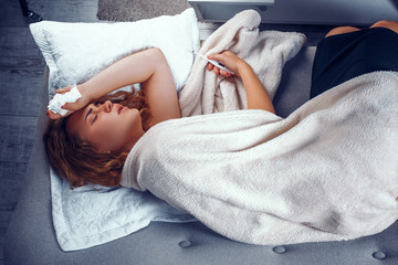 Exhausted woman is laying in bed and holding a thermometer to check temperature. Sick blonde girl laying in bed under wool blanket with a hand with a napkin on her head. - obrazy, fototapety, plakaty