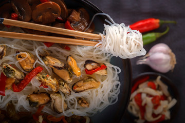 Traditional wooden chopsticks with coiled rice noodles on the background of a dish with seafood, vegetables and peppers. Close-up. Top view