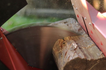 A red safe circular saw sawing an old wooden beam (joist, rafter). The disc of this buzz saw is rotating and cutting the wood for fire. The Sun is shining on a lot of sawdust what flying all around.