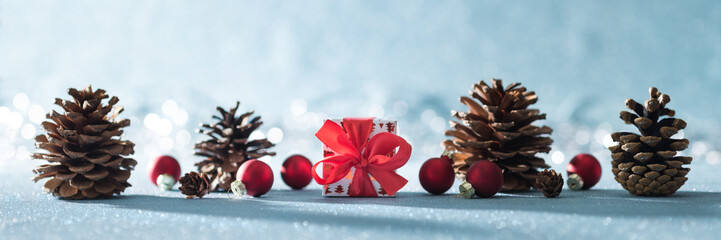 Beautiful simple Christmas banner with copy space. Cute Christmas present, red ornaments and pine cones on shiny blue background with defocused christmas lights.