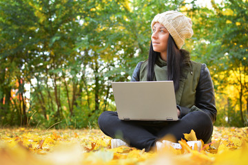 Girl in hipster with laptop in autumn park. A woman in a cap using a laptop while sitting on fallen leaves. Freelancer in the hat uses remote communication technology. Remote work. Free space