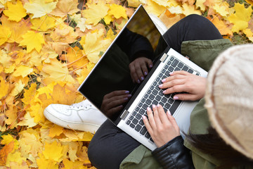 Girl in hipster with laptop in autumn park. A woman in a cap using a laptop while sitting on fallen leaves. Freelancer in the hat uses remote communication technology. Remote work. View from above