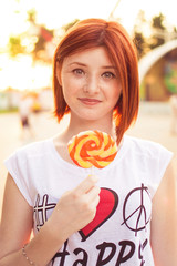 Beautiful young red-haired girl with freckles in funny clothes outdoors at an amusement park is holding a candy on a stick on a summer day. Solar lights up.