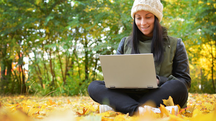 Girl in hipster with laptop in autumn park. A woman in a cap using a laptop while sitting on fallen leaves. Freelancer in the hat uses remote communication technology. Remote work. Free space