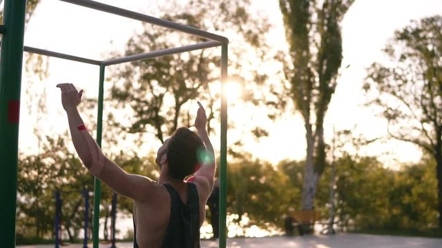 Fit muscular man in black shirt doing pull-ups on horizon bar on sports ground with trees and sun shines on the background. Slow motion