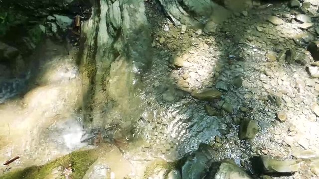 Small creek in the mountains. Pure water flow between old rocks. Moss on the stones, sun flares and shadows around. Sunny summer day. With original sound.
