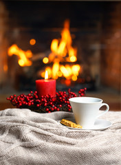 Cup of hot drink with cookie berries and red candle in red Christmas decoration on cozy knitted plaid in front of fireplace. Cozy relaxed magical atmosphere home interior. Christmas New Year concept.