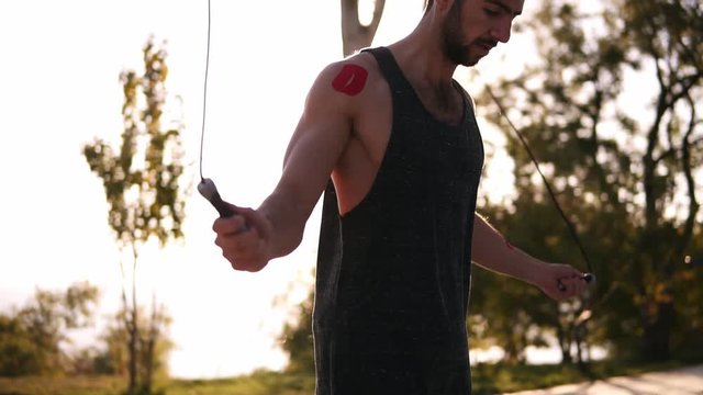Caucasian bearded athlete jumping over skip rope outdoors in the black sportswear. Trees, nature, sun light on the background