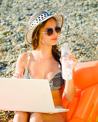Beautiful young woman working with laptop on the tropical beach. Happy traveling woman using laptop on holidays, sitting on yellow inflatable mattress.