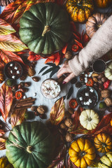 Female hand holding mug with hot chocolate and marshmallows on table with pumpkins and autumn leaves at window. Autumn still life with cozy bokeh lighting. Instagram style