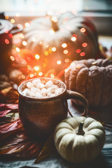 Mug with hot chocolate and marshmallows on table with pumpkins and autumn leaves at window. Autumn...