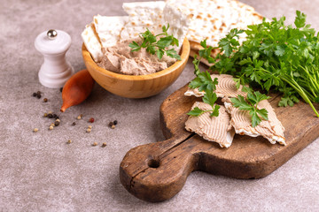 Pieces of matzah with homemade liver pate with parsley and onion on old wooden cutting board with burlap napkin. Gray marble background. Copy space