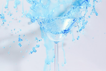 Cocktail with blue liquid in glass. Glass with blue water pouring with liquid with splashes and drops. Martini glass filling with alcohol with splashes on white background. Refreshing drink concept.