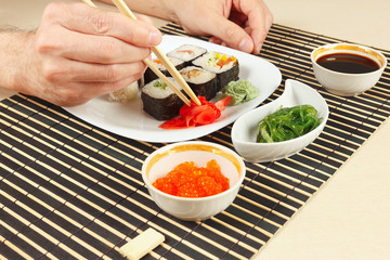 Hands taking ginger with chopsticks. Assorted sushi, hiyashi wakame salad, soy sauce and salmon caviar on a black bamboo mat.