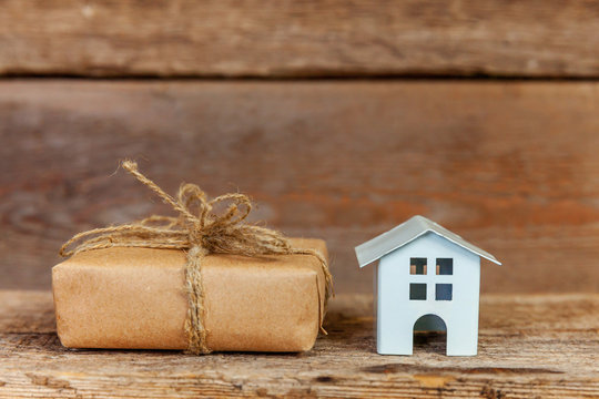 Miniature White Toy House And Gift Box Wrapped Craft Paper On Old Shabby Rustic Wooden Background. Mortgage Property Insurance Dream Home Concept. Buying New House For Family