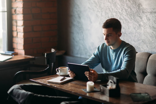 Guy is sitting in a cafe and using a tablet. Student spend time in cafe and drink coffee. He study and note helpful information