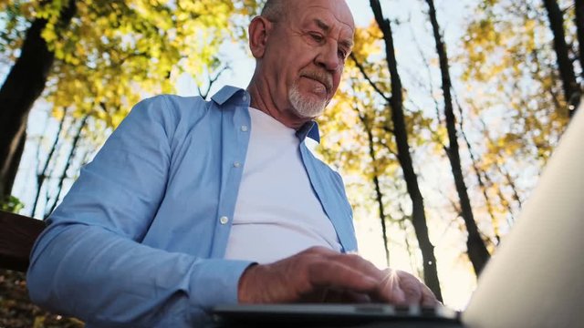 Close up portrait of senior man with a beard using laptop outdoor. Father sits on a park bench and uses hir computer.